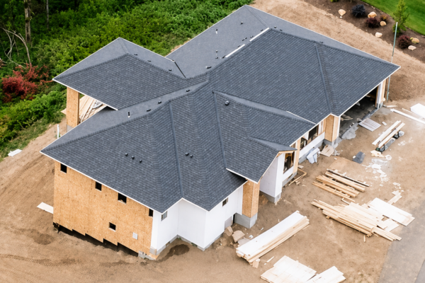 Aerial view of custom home under construction with gray roof, wooded landscape, and building materials on site